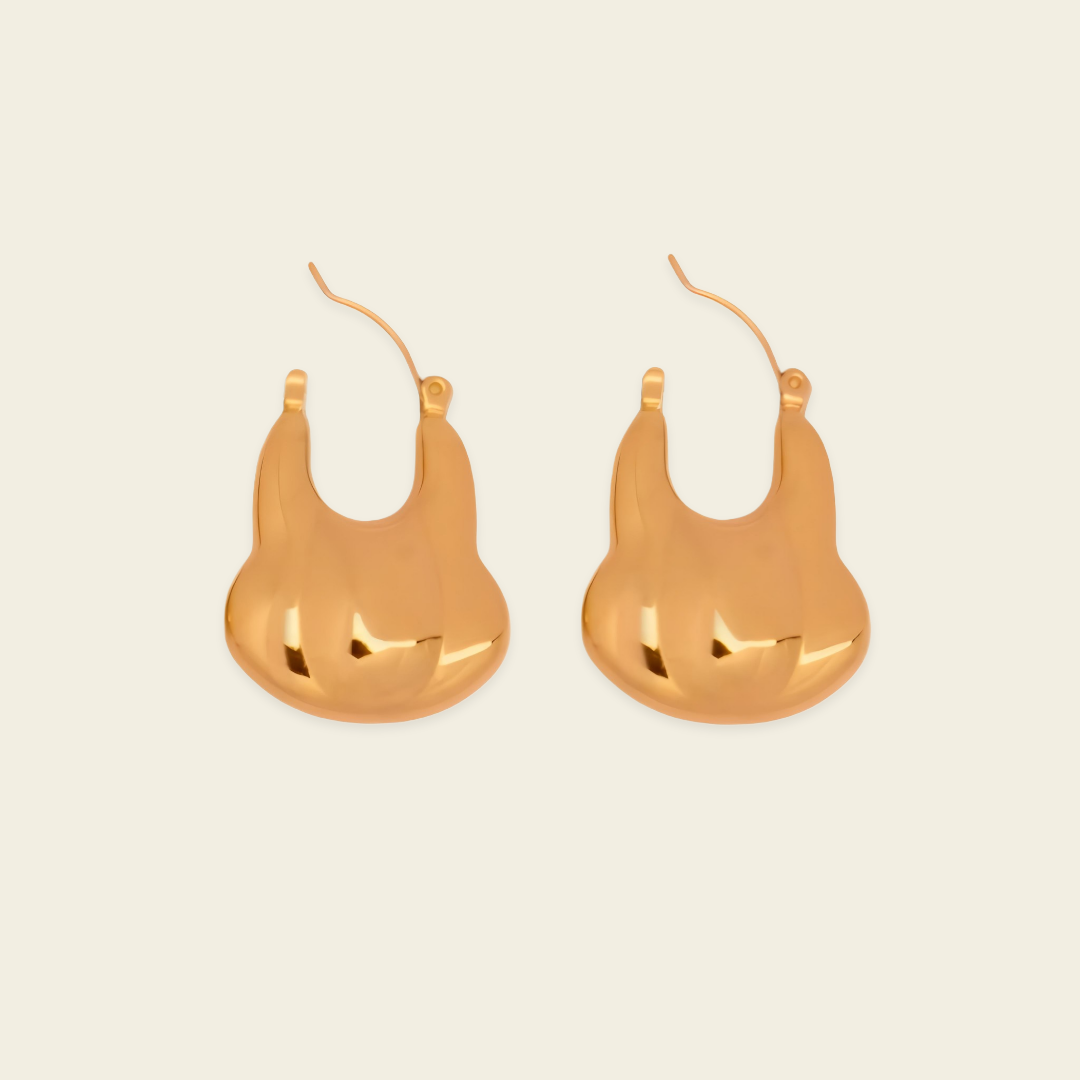 A pair of gold-toned, abstract sculptural hoop earrings on a light cream background.