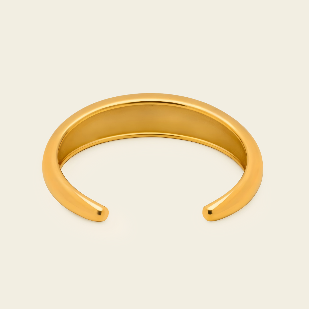 A polished gold open cuff bangle bracelet, photographed on a clean, solid, warm off-white background.