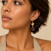 A close-up of a woman wearing a gold drop earring with a small cream-colored stone and a larger purple stone, shown against a warm beige background.