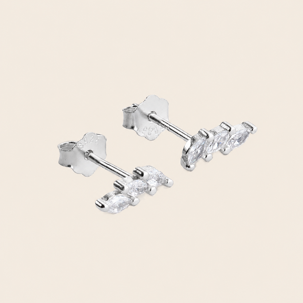 A pair of silver stud earrings with three marquise-cut clear crystals on each earring, displayed on a solid light beige background.