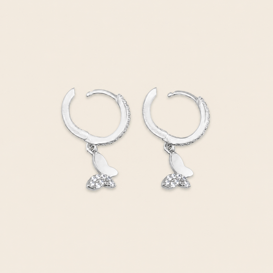 A pair of silver crescent-shaped hoop earrings with dangling butterfly charms, one butterfly in polished silver and the other adorned with small clear crystals, set against a solid light beige background.