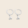 A pair of silver crescent-shaped hoop earrings with dangling butterfly charms, one butterfly in polished silver and the other adorned with small clear crystals, set against a solid light beige background.