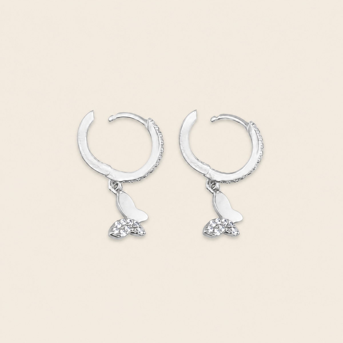 A pair of silver crescent-shaped hoop earrings with dangling butterfly charms, one butterfly in polished silver and the other adorned with small clear crystals, set against a solid light beige background.