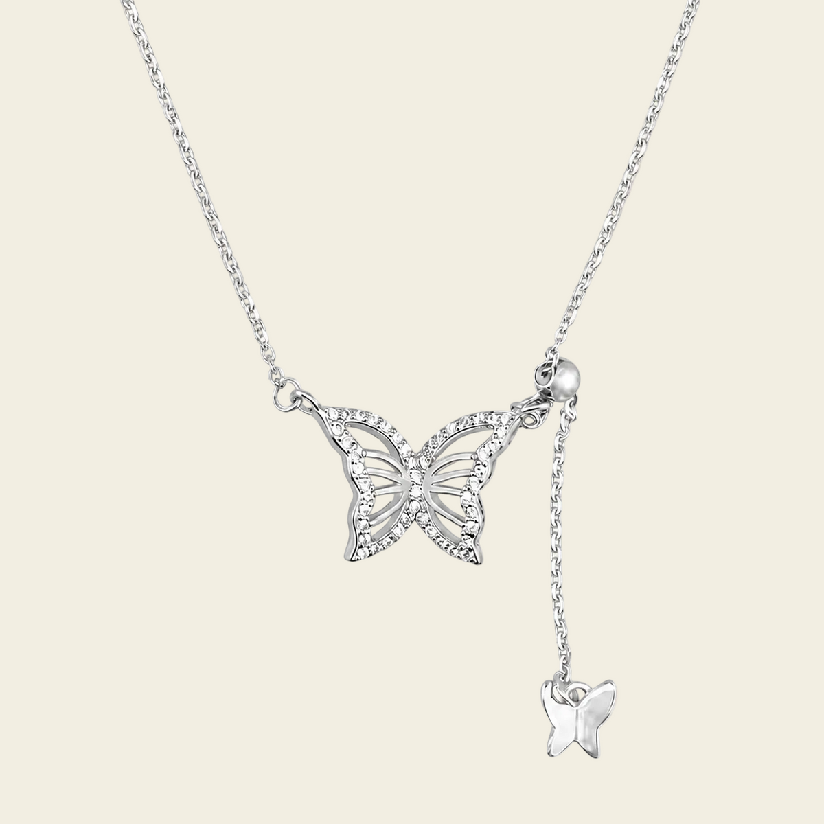 A silver butterfly pendant necklace with a delicate chain, a larger butterfly pendant encrusted with small clear stones, and a smaller dangling butterfly charm, shown on a solid light beige background.