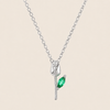A silver pendant necklace featuring a stylized tulip bloom with a marquise-cut green gemstone on the stem, displayed against a solid light beige background.