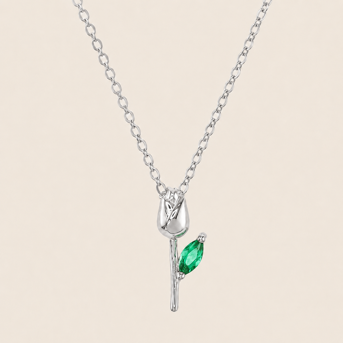 A silver pendant necklace featuring a stylized tulip bloom with a marquise-cut green gemstone on the stem, displayed against a solid light beige background.