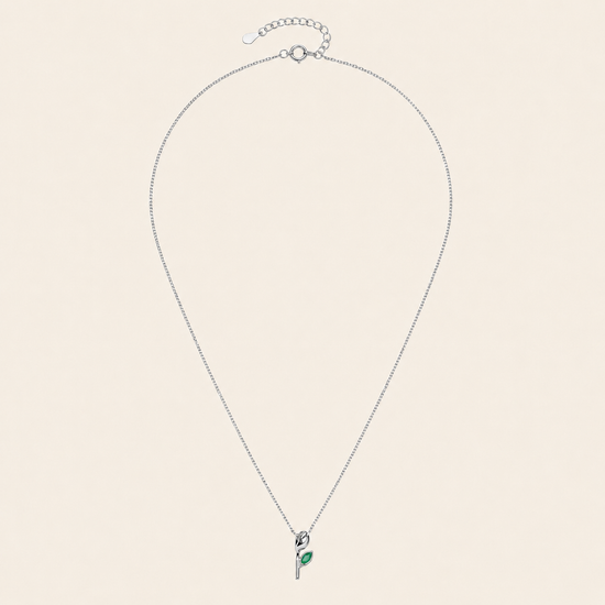A delicate silver chain necklace with a small pendant featuring a green leaf detail, displayed on a solid light beige background.