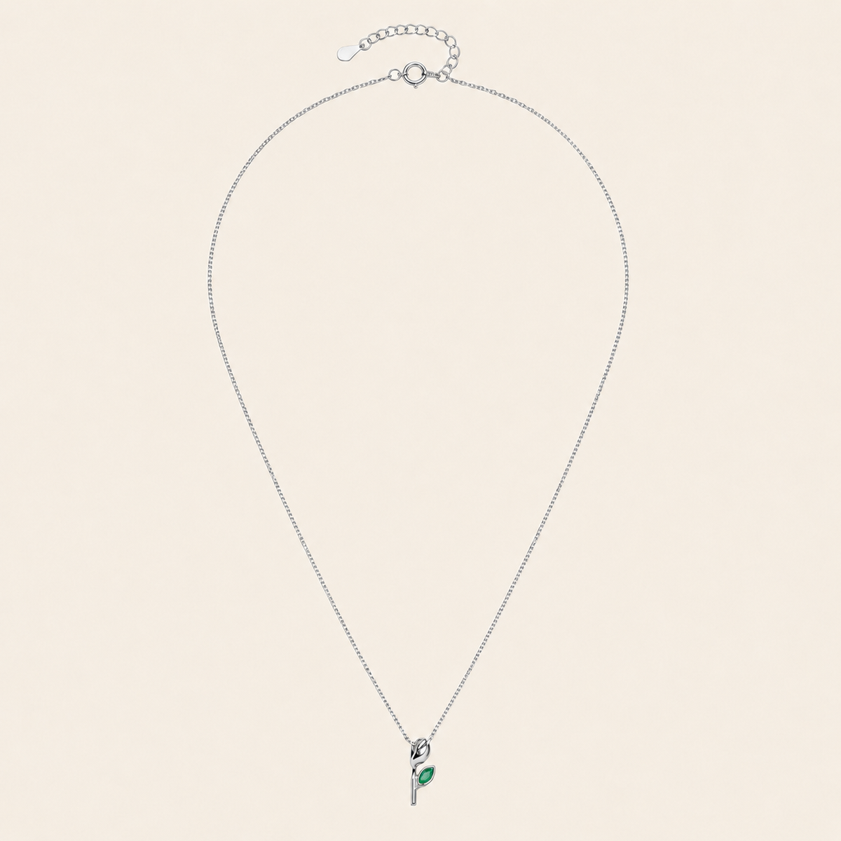 A delicate silver chain necklace with a small pendant featuring a green leaf detail, displayed on a solid light beige background.