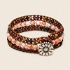 A multi-strand wrap bracelet featuring polished stone beads in earthy brown, cream, and marbled tones, accented by a silver-toned daisy flower charm, set against a solid light beige background.