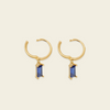 A pair of gold huggie hoop earrings with a small rectangular blue gemstone dangling from the center, set against a solid light beige background.