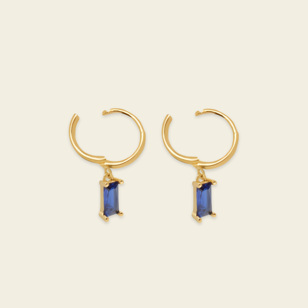 A pair of gold huggie hoop earrings with a small rectangular blue gemstone dangling from the center, set against a solid light beige background.