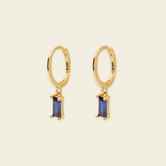 A pair of gold hoop earrings with rectangular blue gemstone drops, shown on a light beige background.