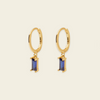 A pair of gold hoop earrings with rectangular blue gemstone drops, shown on a light beige background.
