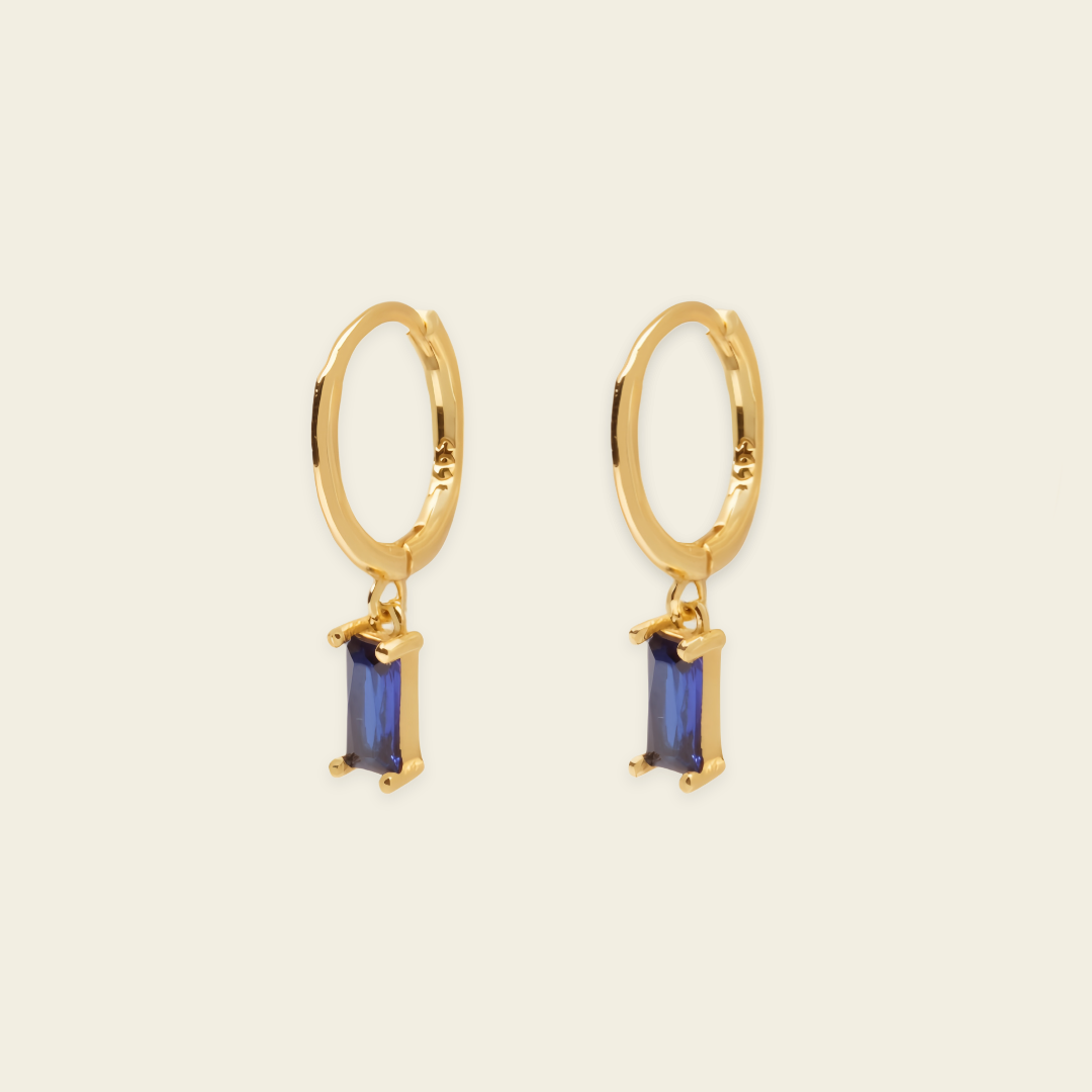 A pair of gold hoop earrings with rectangular blue gemstone drops, shown on a light beige background.