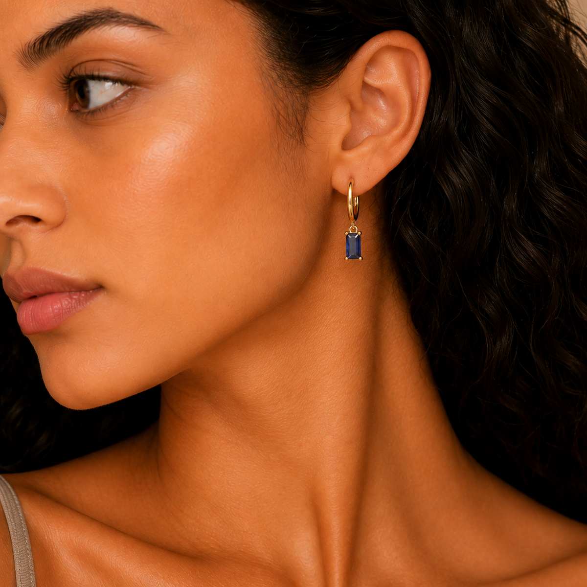 A close-up of a woman wearing a delicate gold huggie hoop earring with a small rectangular blue gemstone dangle, shown against her ear and dark hair.