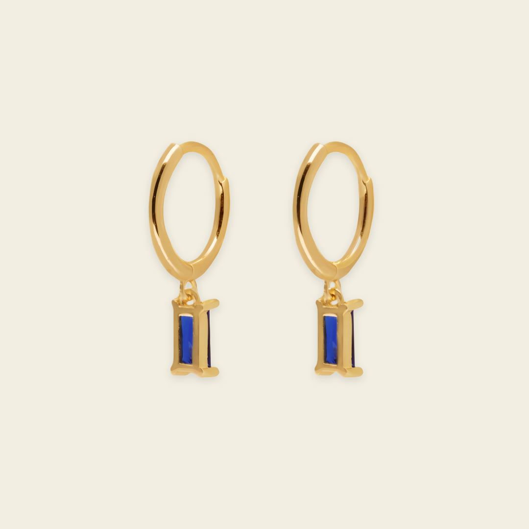 A pair of gold hoop earrings with a small rectangular blue enamel charm, displayed on a light cream background.