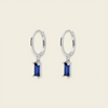 A pair of gold hoop earrings with rectangular blue gemstone drops, shown on a light beige background.