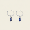 A pair of huggie hoop earrings with a small rectangular blue gemstone dangling from the center, set against a solid light beige background.