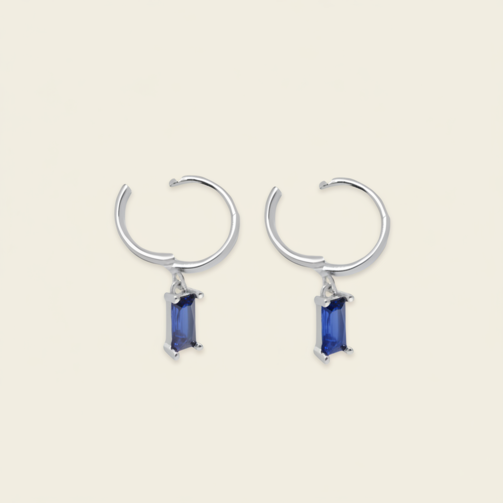 A pair of huggie hoop earrings with a small rectangular blue gemstone dangling from the center, set against a solid light beige background.