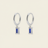 A pair of gold hoop earrings with a small rectangular blue enamel charm, displayed on a light cream background.
