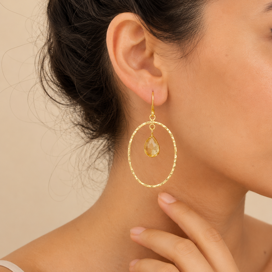 Gold hoop earrings with a teardrop-shaped gemstone worn by a person against a beige background