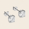 A pair of round-cut diamond stud earrings in a silver four-prong setting, shown on a light beige background.