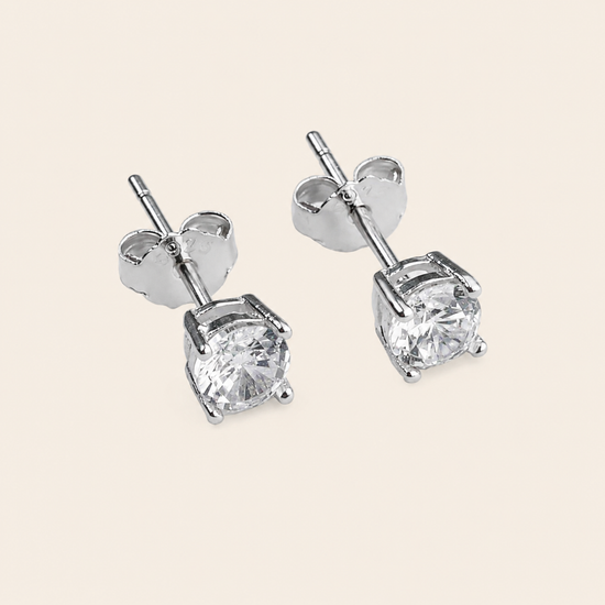 A pair of silver stud earrings with round clear gemstones in a four-prong setting, shown on a light beige background.