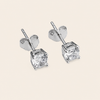 A pair of silver stud earrings with round clear gemstones in a four-prong setting, shown on a light beige background.