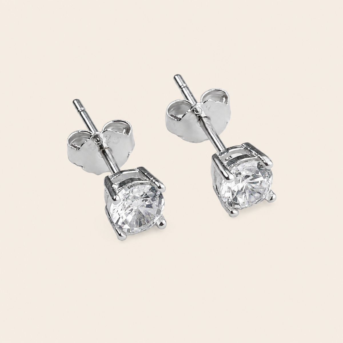 A pair of silver stud earrings with round clear gemstones in a four-prong setting, shown on a light beige background.