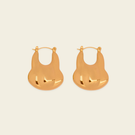 A pair of gold-toned, organic-shaped hoop earrings with a polished, reflective finish, displayed on a solid light beige background.