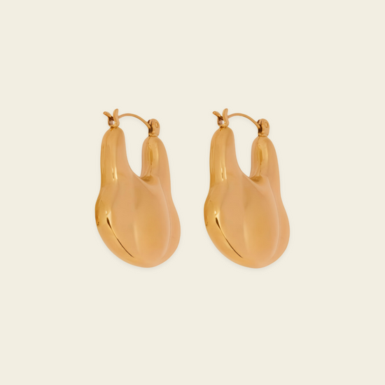 A pair of gold-toned, organic-shaped hoop earrings with a polished finish, displayed on a solid light beige background.