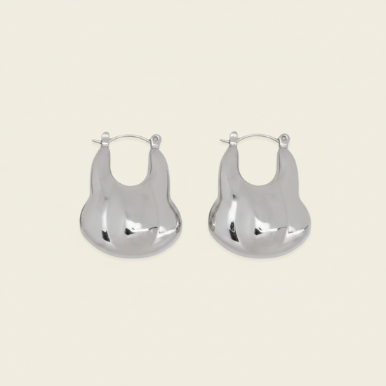 A pair of silver-toned, organic-shaped hoop earrings with a polished, reflective finish, displayed on a solid light beige background.