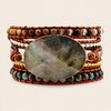 A multi-strand wrap bracelet featuring a large oval labradorite stone centerpiece in a gold bezel, surrounded by brown leather cords, multi-colored beads, and a gold chain, set against a light beige background.