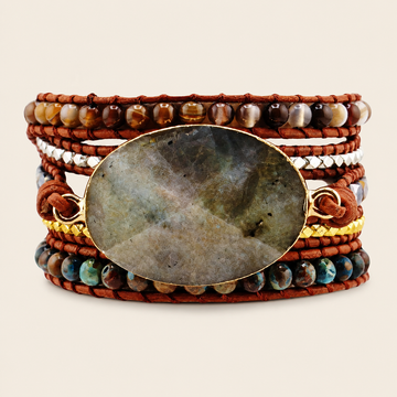 A multi-strand wrap bracelet featuring a large oval labradorite stone centerpiece in a gold bezel, surrounded by brown leather cords, multi-colored beads, and a gold chain, set against a light beige background.