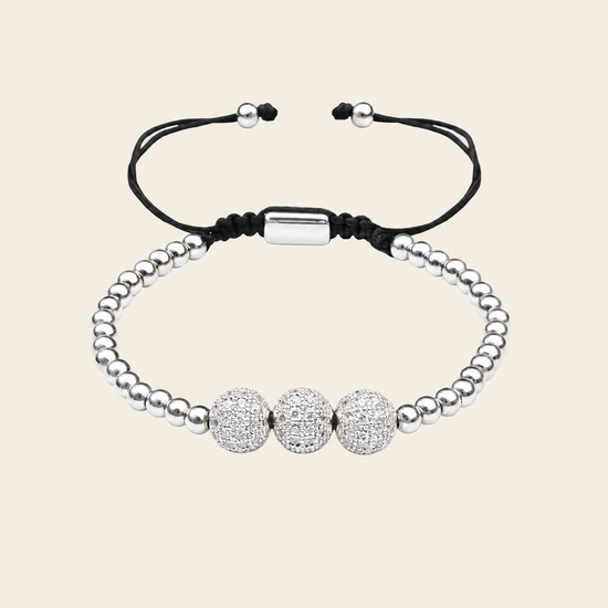 A silver beaded bracelet with three round pavé-style center beads and adjustable black cord ends, shown on a plain warm beige background.