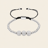 A silver beaded bracelet with three round pavé-style center beads and adjustable black cord ends, shown on a plain warm beige background.
