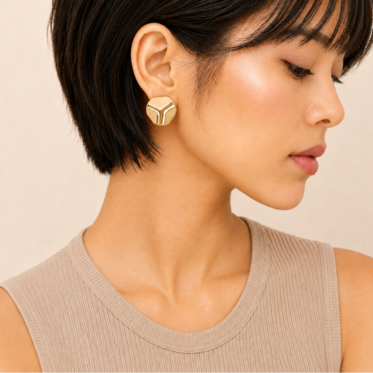A close-up of a woman with short dark hair wearing a geometric gold earring with a white chevron pattern, paired with a beige ribbed tank top against a soft beige background.