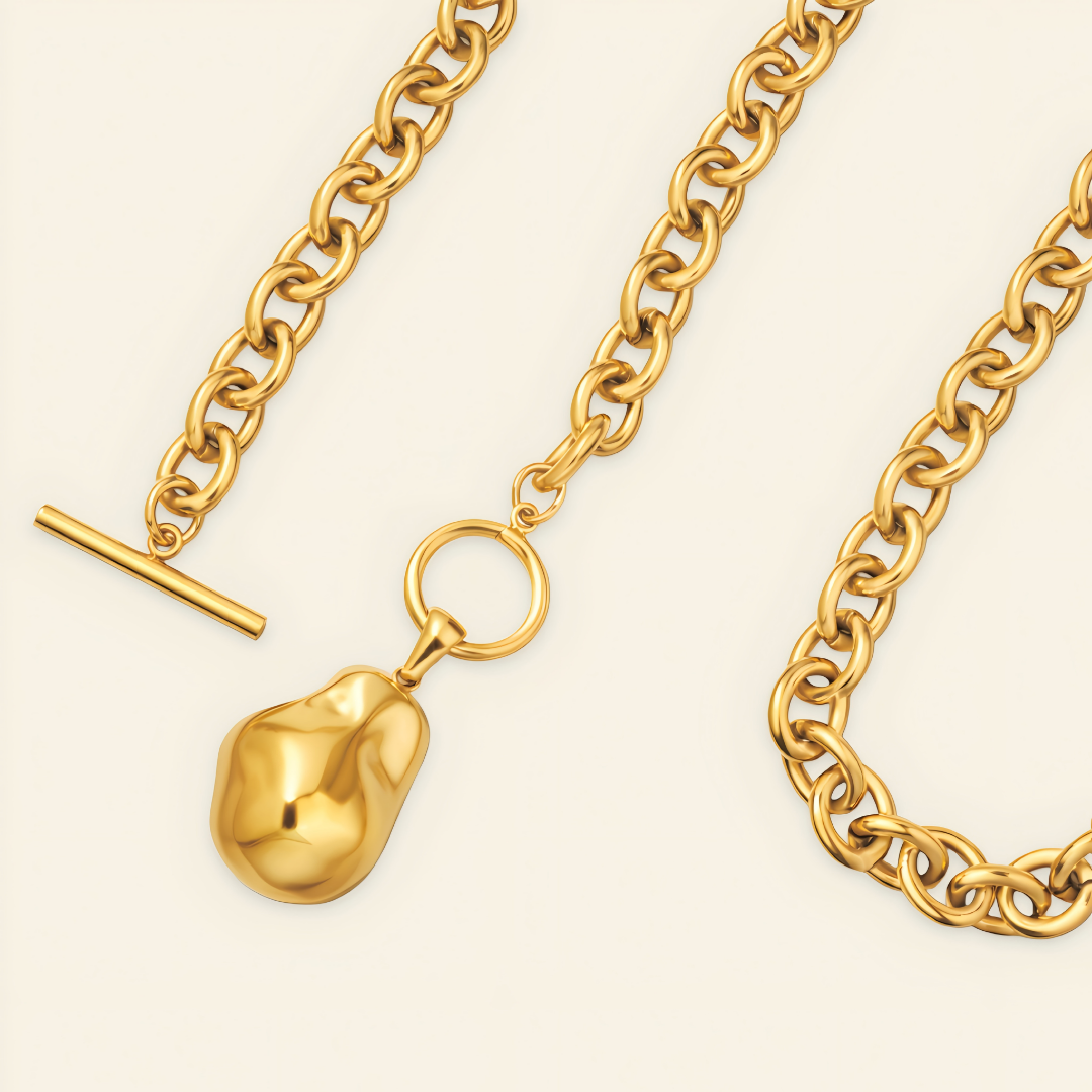 Gold chain necklace with a large irregular pendant, a ring connector, and a T-bar clasp, shown on a light cream background.