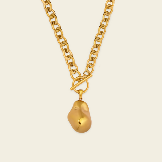 A gold chain necklace with a toggle clasp and an irregular, organic-shaped gold pendant, shown against a solid light beige background.