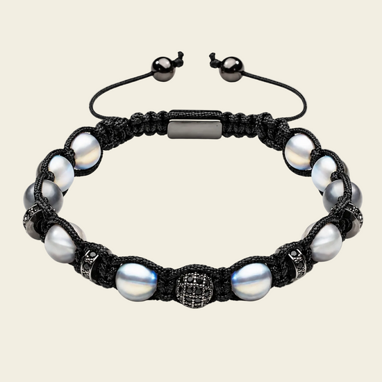 A black macrame bracelet featuring large iridescent white pearls, a textured silver-toned central bead, and a rectangular silver-toned clasp, shown on a light beige background.