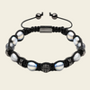 A black macrame bracelet featuring large iridescent white pearls, a textured silver-toned central bead, and a rectangular silver-toned clasp, shown on a light beige background.