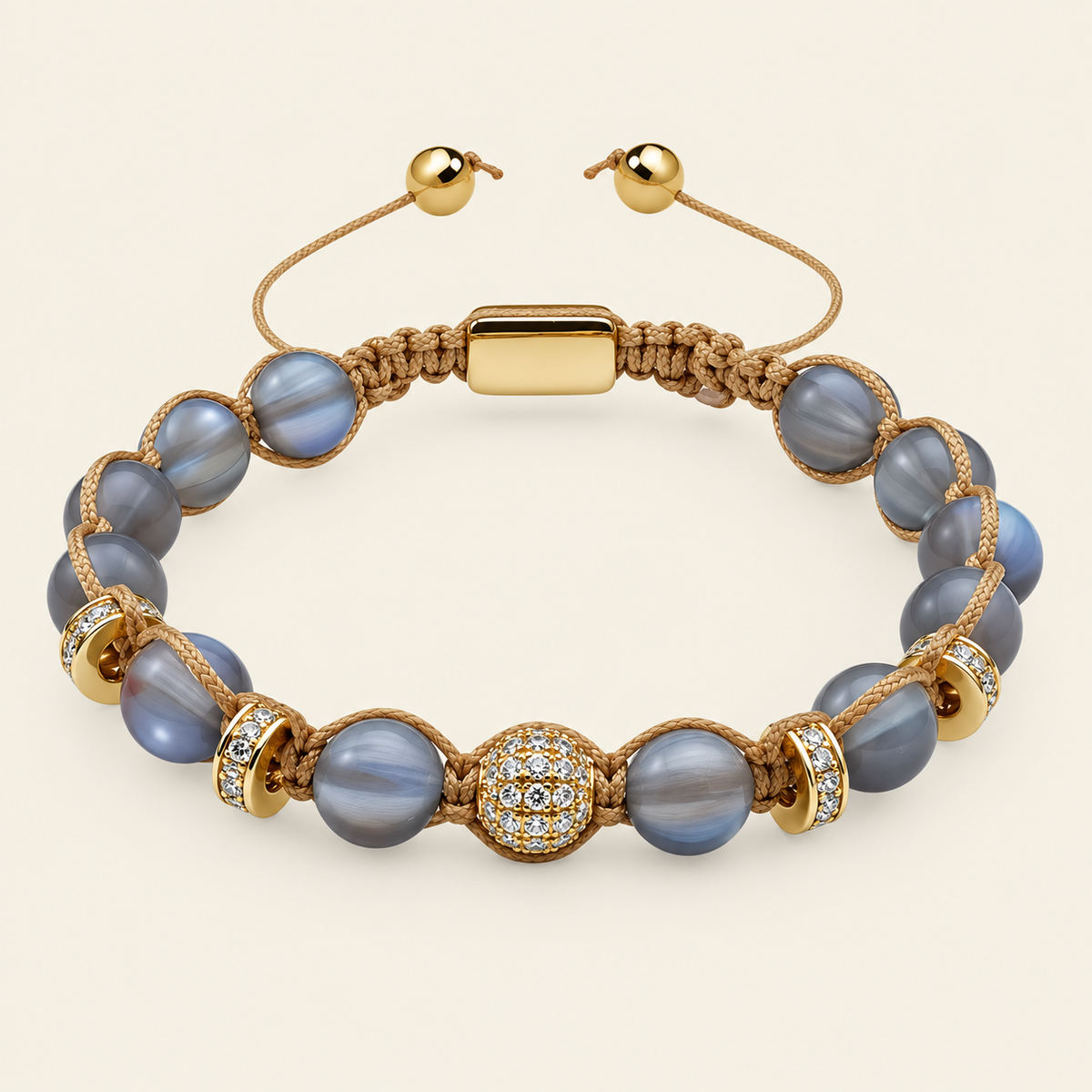 A gold-plated beaded bracelet with round blue-gray beads, a central pavé detail, and a gold-plated clasp, shown on a light beige background.