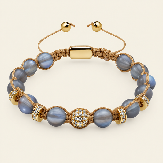 A gold-plated beaded bracelet with round blue-gray beads, a central pavé detail, and a gold-plated clasp, shown on a light beige background.