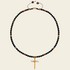 A beaded necklace featuring small black beads interspersed with metallic gold and brown beads, with a gold crucifix pendant at the center and an adjustable sliding cord closure at the top, shown against a solid light beige background.