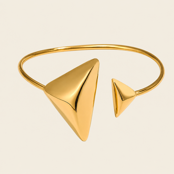 A polished gold-tone open cuff bangle bracelet featuring two triangular pyramid-like geometric accents, shown on a plain light beige background.