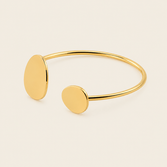 A minimalist gold open cuff bracelet with two flat oval disc charms, photographed on a clean, light beige background.