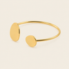 A minimalist gold open cuff bracelet with two flat oval disc charms, photographed on a clean, light beige background.