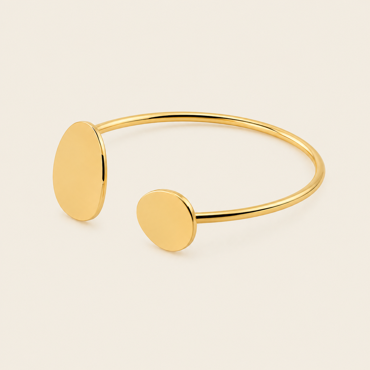 A minimalist gold open cuff bracelet with two flat oval disc charms, photographed on a clean, light beige background.