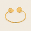 A polished gold-tone open cuff bracelet with two flat circular disc ends, displayed on a solid light beige background.