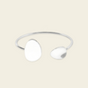 A minimalist silver-toned open cuff bangle featuring two smooth, organic-shaped disc elements, displayed on a solid light beige background.